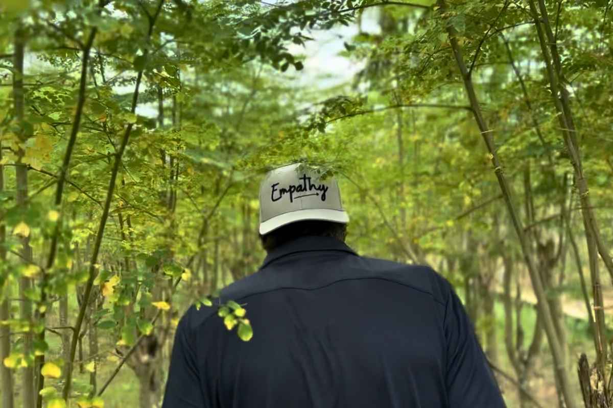 A person viewed from behind wearing a dark navy shirt and a white trucker-style cap with the word 'Empathy' printed in black script, standing in a wooded area with green trees and bamboo.